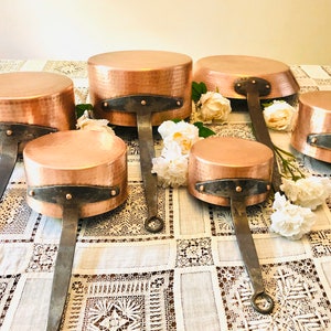 May include: Set of six copper saucepans with hammered surfaces and iron handles. The saucepans are arranged in a staggered pattern on a white lace tablecloth.