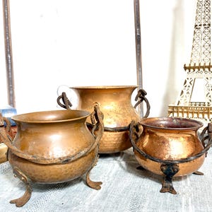 May include: Three antique copper cauldrons with hammered textures and dark metal handles and feet. The cauldrons vary in size, with the largest in the background and the smallest in the foreground. The copper has a warm, aged patina.