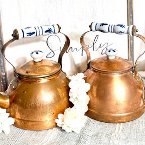 May include: Two copper tea kettles with rounded bodies and curved spouts. Each kettle features a white ceramic handle with blue floral patterns and a matching white ceramic knob on the lid. The kettles are positioned side by side.