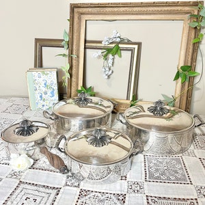 May include: Four silver serving dishes with lids and ornate handles. The dishes are arranged on a white lace tablecloth with a floral arrangement in the background.