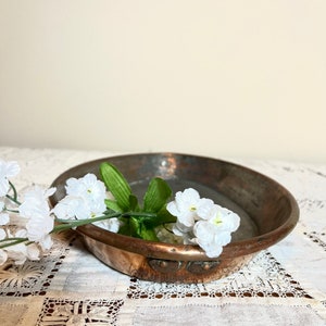 May include: A round copper serving dish with a slightly hammered finish. The dish has two decorative handles on the side and is sitting on a white lace tablecloth with white flowers.