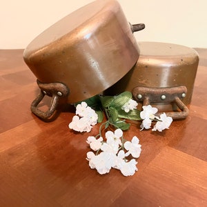 May include: Two vintage copper pots with worn handles, stacked on a wooden surface with white flowers.