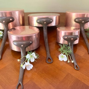 May include: Set of five copper saucepans with black handles. The saucepans are arranged in a circle on a wooden surface. The saucepans are different sizes and have a shiny copper finish.