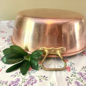 May include: A large copper pot with a brass handle. The pot is shiny and has a rounded bottom. It is sitting on a white tablecloth with a floral pattern.