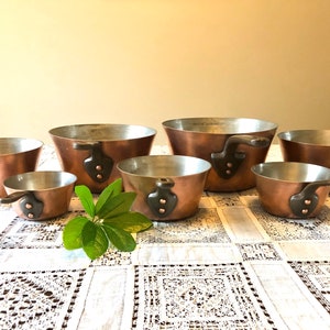 May include: Set of six copper saucepans with black handles and copper rivets. The saucepans are arranged in size order from largest to smallest.