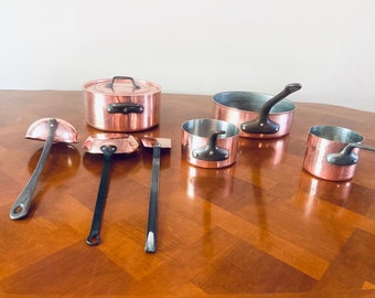 May include: A set of copper cookware with black handles, including a pot with a lid, a ladle, a small saucepan, and two smaller saucepans. The cookware is arranged on a wooden table.