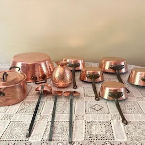 May include: A set of copper cookware, including a large pot with a lid, a smaller pot, a pitcher, and several saucepans. The cookware is all hammered and has a shiny finish. The cookware is arranged on a white tablecloth with a lace pattern.