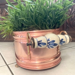 May include: Copper planter with a blue and white ceramic handle. The planter is oval shaped and has a decorative rim. The planter is filled with green plants.
