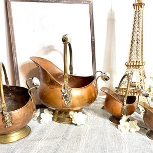 May include: A set of four graduated copper coal scuttles with brass handles, pedestal bases, and decorative metal accents. Each scuttle features a curved spout, a swing handle with a white ceramic grip, and a polished finish on the exterior.