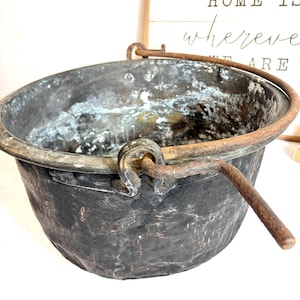 Peut inclure: Un chaudron antique en métal noir avec une anse incurvée. Le chaudron présente un aspect vieilli et rustique, avec des signes d'usure visibles. La poignée est en métal similaire et est fixée au bord. En arrière-plan, une enseigne en bois avec le texte "HOME IS where we ARE".