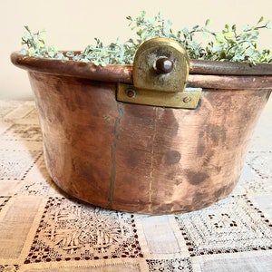 May include: A large copper pot with a brass handle and a decorative brass knob. The pot is filled with green foliage and is sitting on a white lace tablecloth.
