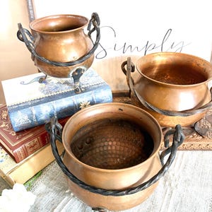 May include: Three antique copper cauldrons with twisted iron handles and decorative feet. The cauldrons are displayed on a table with books and a floral arrangement. The copper has a warm, aged patina.
