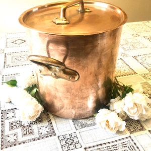 Peut inclure: Une grande casserole en cuivre avec un couvercle et une seule poignée. La casserole est posée sur une nappe en dentelle blanche avec des fleurs blanches.