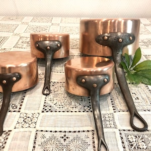 May include: Set of four copper saucepans with black handles. The saucepans are nested together on a white lace tablecloth. The saucepans have numbers on the bottom, indicating their size.