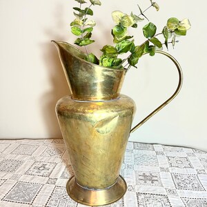 May include: A tall, gold-toned brass pitcher with a hammered finish and a wide, curved spout. The pitcher has a single handle and is filled with green eucalyptus stems.