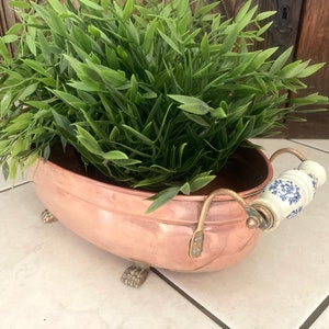 May include: A copper planter with a white and blue porcelain handle. The planter is oval shaped and has four decorative feet. The planter is filled with green leafy plants.