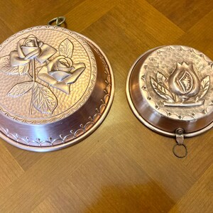 May include: Two antique copper cake molds with decorative floral designs. The larger mold features two roses with leaves, while the smaller mold has a single stylized flower.