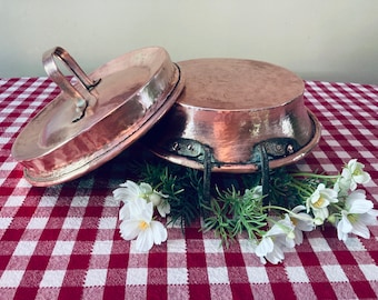 May include: Two antique copper pots with handles. The larger pot has three iron handles and the smaller pot has one copper handle. The pots are sitting on a red and white checkered tablecloth with white flowers.