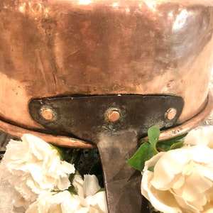 May include: A vintage copper pot with a dark brown iron handle and three rivets. The pot has a worn, aged patina and is sitting on a white lace tablecloth.
