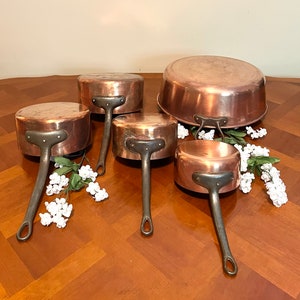 May include: A set of five copper saucepans with black handles. The saucepans are arranged in size order, from largest to smallest. The largest saucepan is a wide-based, rounded-bottomed pot. The other four saucepans are smaller and have a more traditional shape. All the saucepans are made of copper and have a shiny finish. The handles are made of black metal and are attached to the saucepans with rivets.