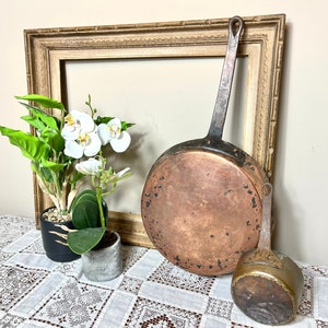 May include: Two copper pots, one large and one small, with handles. The large pot is hanging from a wooden frame. A potted plant with white flowers is in the foreground.