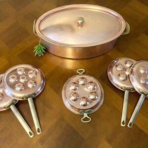 May include: Set of copper cookware, including a large oval pot with a lid and four smaller round pans with handles. The pans have a unique design with multiple indentations on the bottom.