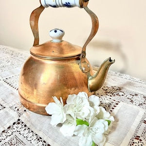 May include: A copper teapot with a white and blue porcelain handle and lid. The teapot is sitting on a white lace tablecloth with white flowers.