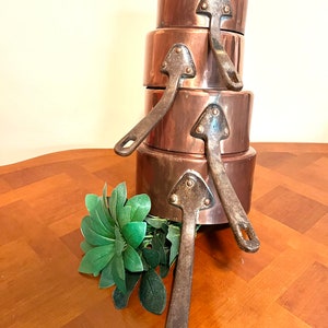 May include: A set of four copper pots with black iron handles. The pots are stacked on top of each other, with the smallest pot on top. The pots are all in good condition and have a shiny finish.