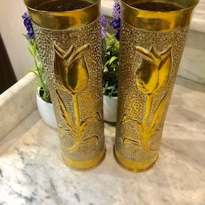 May include: Two gold metal vases with a textured surface and a raised tulip design. The vases are cylindrical and have a wide opening at the top.