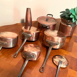 May include: A set of six copper pots with long handles. The pots are of varying sizes and are arranged on a wooden table.