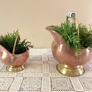 May include: Two copper and brass coal scuttles with handles, filled with greenery, sit on a lace tablecloth. The scuttles have a copper body and brass base. The handles have white ceramic ends with blue floral designs.