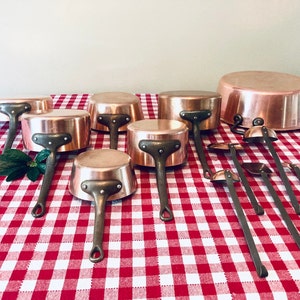 May include: A set of copper pots and ladles on a red and white checkered tablecloth. The pots are various sizes and have copper handles. The ladles are also copper and have long wooden handles.