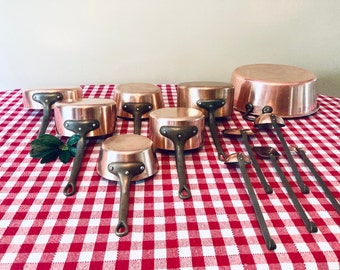 May include: A set of copper pots and ladles on a red and white checkered tablecloth. The pots are various sizes and have copper handles. The ladles are also copper and have long wooden handles.