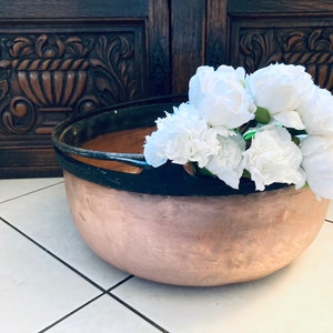 May include: A copper pot with a black rim and handle, filled with a bouquet of white flowers.