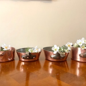 May include: Four copper saucepans with black handles, each filled with white flowers.