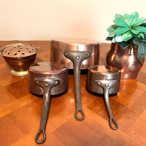May include: Set of four copper pots with black handles. The pots are of varying sizes and are arranged on a wooden table. A small copper pot with holes is in the center of the table. A copper vase with a green plant is on the right side of the table.