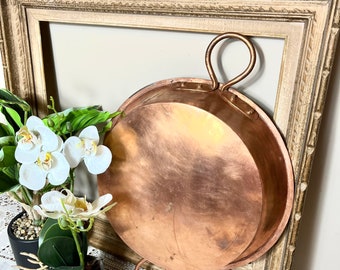 May include: A large, round copper pan with two loop handles. The pan is hanging on a wooden picture frame.