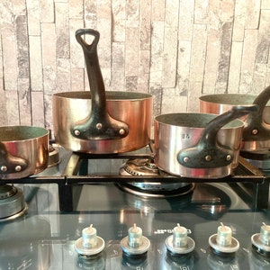 May include: Four copper pots with black handles sit on a black stovetop. The pots are different sizes and are arranged in a row. The stovetop has four silver knobs.