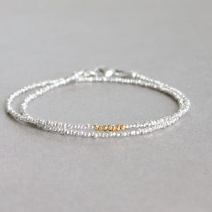 May include: A delicate silver beaded bracelet with a double strand design. The bracelet features a small section of gold-colored beads in the center. The clasp is silver. The jewelry is displayed on a light gray surface.