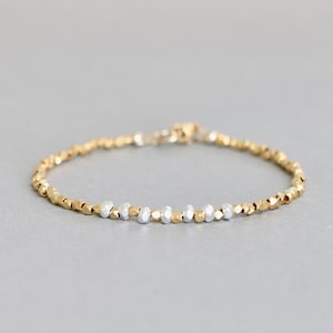 May include: A delicate bracelet featuring small, faceted beads in gold and silver tones. The bracelet is strung together, creating a flexible band. The beads have a textured appearance, reflecting light. The jewelry is displayed on a neutral gray surface.