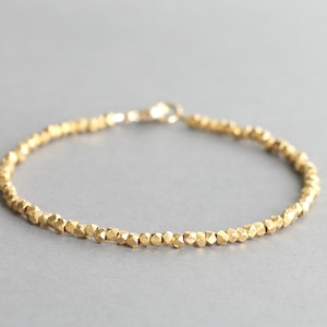 May include: A delicate gold bracelet composed of numerous small, irregularly shaped, faceted beads that create a textured, shimmering appearance. The bracelet features a simple circular clasp mechanism at the end of the chain.