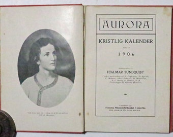 May include: A vintage book cover for the 1906 Aurora Kristlig Kalender, a Christian calendar published in Swedish. The cover features a black and white portrait of a young woman with a serious expression. The title "Aurora Kristlig Kalender" is printed in bold, black letters at the top of the cover. The year "1906" is printed below the title.