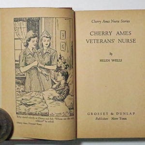 May include: An open book titled "Cherry Ames Veterans' Nurse" by Helen Wells. The book's illustration depicts two nurses attending to a young patient in a hospital bed. The book is open to a page with text and an illustration.