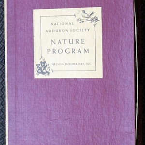 Peut inclure: Un livre vintage avec une couverture violet foncé texturée. Le titre "Nature Program" est affiché sur une étiquette crème, avec le texte "National Audubon Society" et "Nelson Doubleday, Inc.". Des illustrations décoratives d'un oiseau et de feuillage sont également présentes.