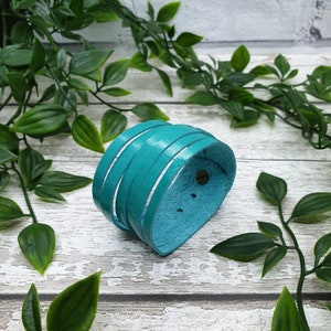 May include: A teal leather cuff bracelet with several horizontal straps. The bracelet is curved and has a metal stud fastening. The leather has a smooth, slightly shiny finish. The background includes green leaves and a light-coloured wooden surface.