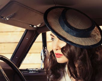 Puede incluir: Una mujer con un sombrero de paja de ala ancha con una banda azul, sentada en un coche antiguo. El sombrero está inclinado hacia abajo, cubriendo parcialmente su rostro. Ella está mirando por la ventana.
