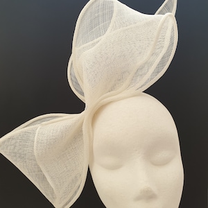 May include: A white, delicate, and intricately woven hat with a large bow. The hat is made of a soft, airy material and has a unique, asymmetrical design.