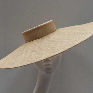 Puede incluir: Un sombrero de ala ancha hecho de paja en un color beige claro. El sombrero tiene una copa plana y un ala ancha y ligeramente curvada. El ala está ligeramente inclinada hacia arriba en un lado.