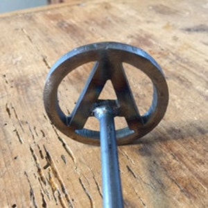 Op de afbeelding: Een metalen brandmerk met een ronde kop met de letter "A" in een gestileerd lettertype. Het merk heeft een lange, dunne steel.