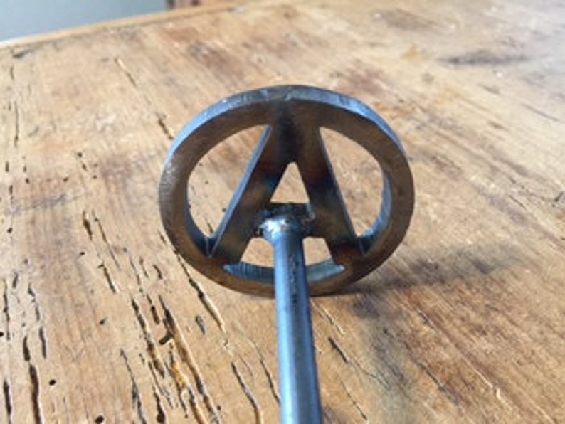 Op de afbeelding: Een metalen brandmerk met een ronde kop met de letter "A" in een gestileerd lettertype. Het merk heeft een lange, dunne steel.
